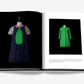 Open book displaying two fashion garments on mannequins: a short-sleeve mini dress with a large green patch and a long-sleeve green dress, both against a black background. Fashion enthusiasts will appreciate the haute couture vibe reminiscent of Dior: Raf Simons.