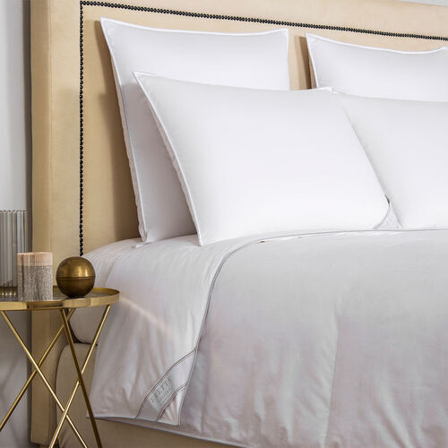 A neatly made bed with 380 thread count cotton sateen white pillows and a Frette Cortina Duvet Insert, Light Density next to a round side table with candles and a decorative gold sphere.