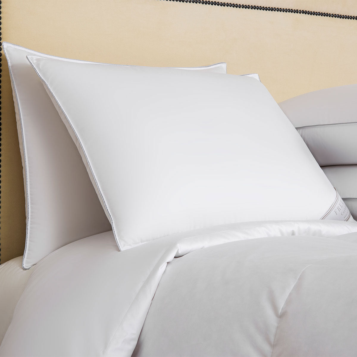 Two white Frette Cortina Pillow Inserts, Firm Density, filled with Polish goose down and covered in smooth, clean pillowcases are placed on a neatly made bed with white bedding. The beige headboard is partially visible in the background.