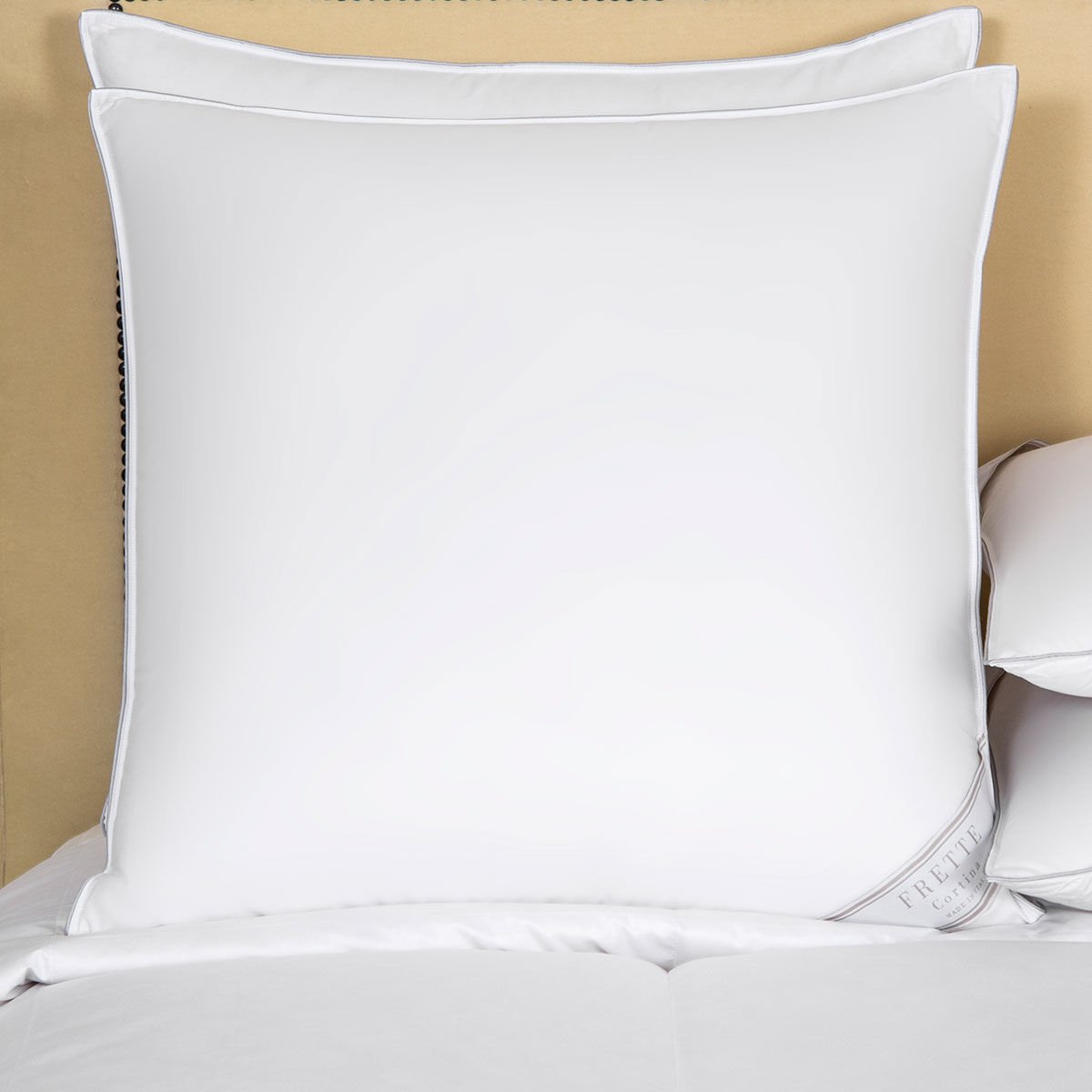 A large, fluffy Frette Cortina Pillow Insert, Firm Density on a neatly made bed partially covers another smaller pillow. The bed, adorned with white linens, exudes comfort and luxury with its cotton sateen finish.