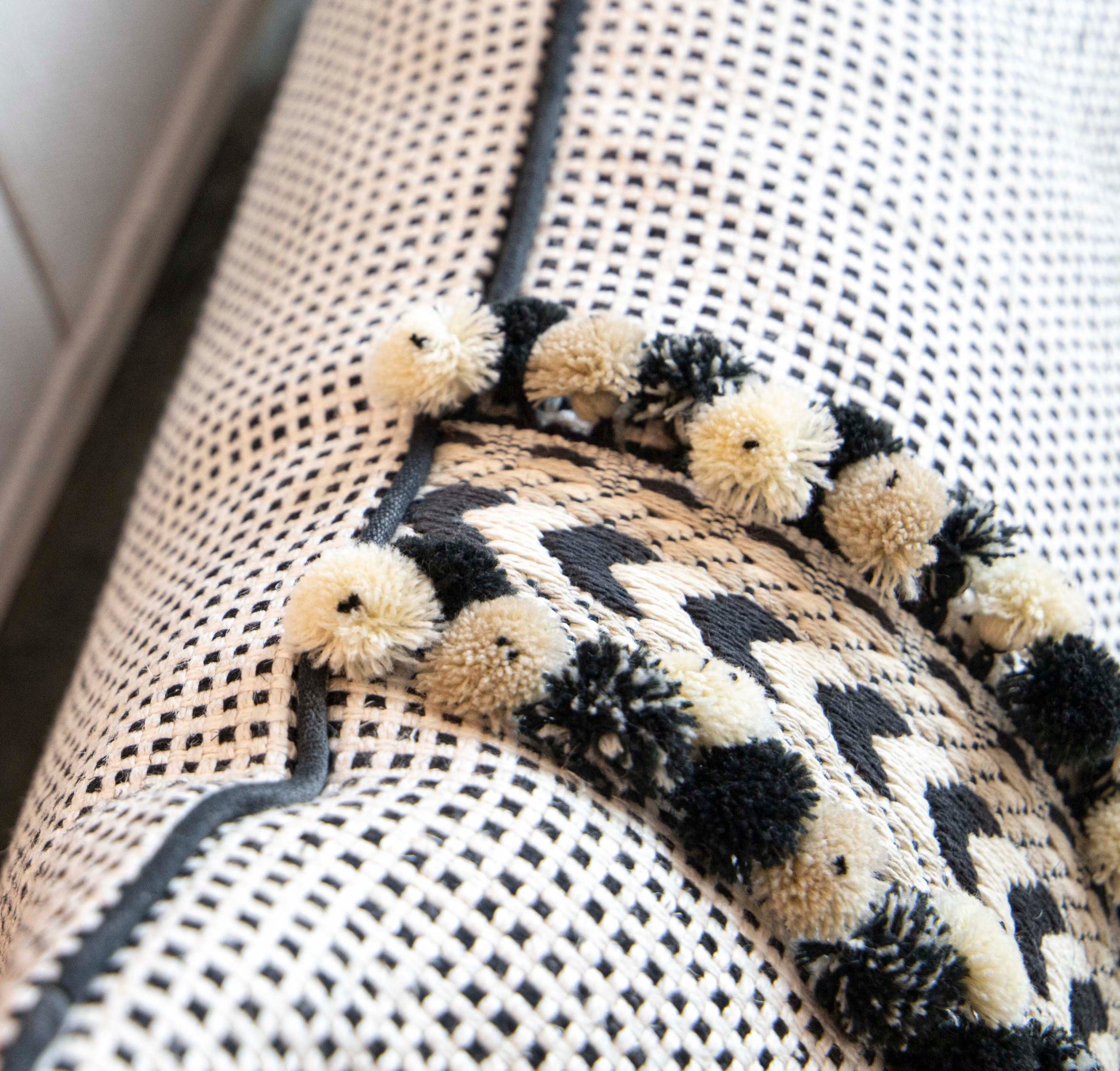 Close-up of a textured fabric with a checkered pattern and a black and white zigzag design. Kumano Weave Maracana Pom Stripe Pillow using the finest materials are adorned with small pom-poms along the edge, showcasing special attention to detail.