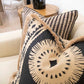 Black and beige Bora Bora Lava Black Pillow, custom-made from the finest materials, boast intricate patterns and fringed edges as they adorn a light-colored sofa. In the background, a side table with a lamp and other decor items completes the elegant setting.