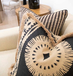 Black and beige Bora Bora Lava Black Pillow, custom-made from the finest materials, boast intricate patterns and fringed edges as they adorn a light-colored sofa. In the background, a side table with a lamp and other decor items completes the elegant setting.