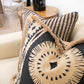 Baleen Pillows with geometric patterns and fringe detailing, including double sided pillows, are placed on a beige sofa. An end table with a black vase and additional seating are in the background.