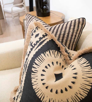 Baleen Pillows with geometric patterns and fringe detailing, including double sided pillows, are placed on a beige sofa. An end table with a black vase and additional seating are in the background.