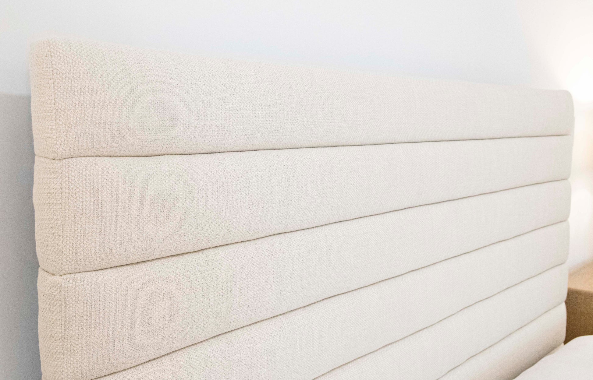 Close-up of a beige, upholstered Channel Bed, King headboard with horizontal stitching lines and elegant channeled detailing, set against a light-colored wall.