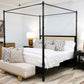 A bedroom with a black Willa Raffia Bed, King featuring turned bedposts, striped and patterned pillows, bedside tables with lamps, a bench at the foot of the bed, and a dresser with books in the background.