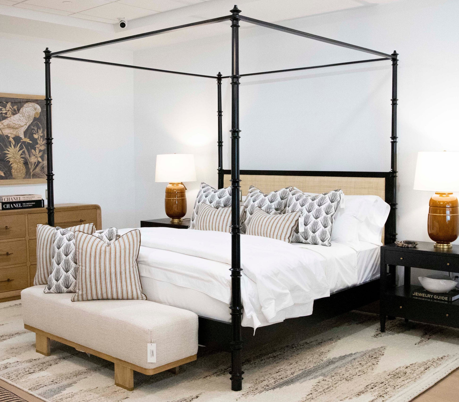 A bedroom with a black Willa Raffia Bed, King featuring turned bedposts, striped and patterned pillows, bedside tables with lamps, a bench at the foot of the bed, and a dresser with books in the background.