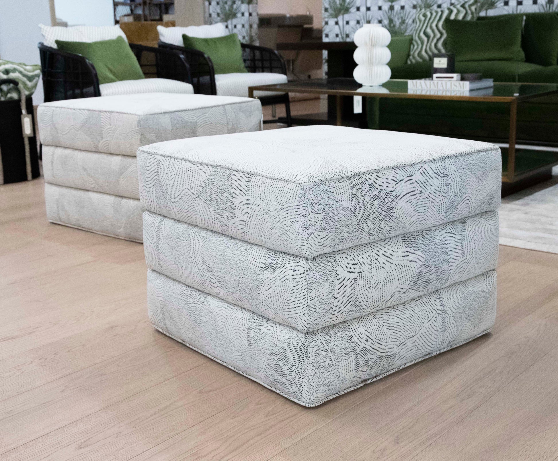 Two light-colored, custom upholstered Channeled Ottomans with a textured pattern are placed on a wooden floor in a living room setting with a green sofa, glass coffee table, and decorative items in the background.