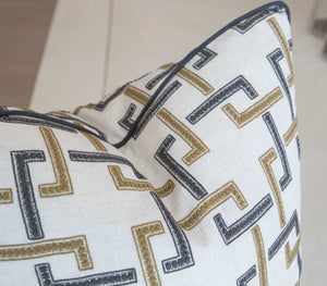 Close-up of a Sekai Charcoal Cinnamon Pillow with a geometric zigzag pattern in gold, black, and gray on a cream background. Crafted with special attention to detail and the finest materials, the pillow features elegant black piping along the edges.