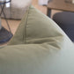 Close-up of a green triangular-shaped cushion on a sofa, showcasing the finest materials. The background is blurred, featuring a striped fabric item and part of a wooden table, highlighting the special attention to detail in this Lucido D'albano Pillow.