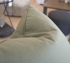 Close-up of a green triangular-shaped cushion on a sofa, showcasing the finest materials. The background is blurred, featuring a striped fabric item and part of a wooden table, highlighting the special attention to detail in this Lucido D'albano Pillow.