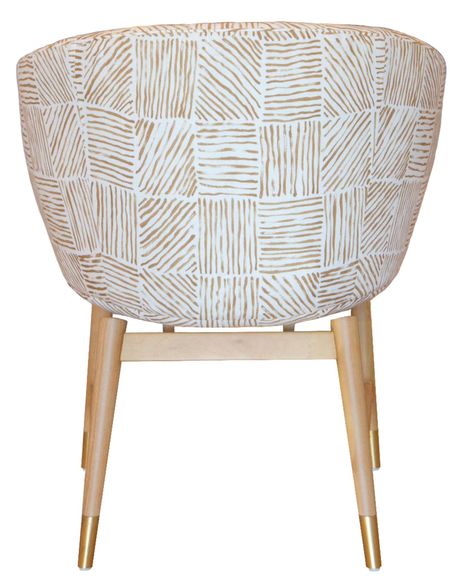 The Coupe Chair features a modern design with white and gold geometric-patterned fabric. Its mid-century style is complemented by light wooden legs adorned with gold accents at the tips.