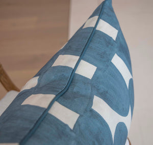 Close-up view of an Arch Sea Pillow with white geometric patterns, crafted from the finest materials, showing the seam and fabric texture. The background includes a small portion of a wooden chair and a light-colored floor.