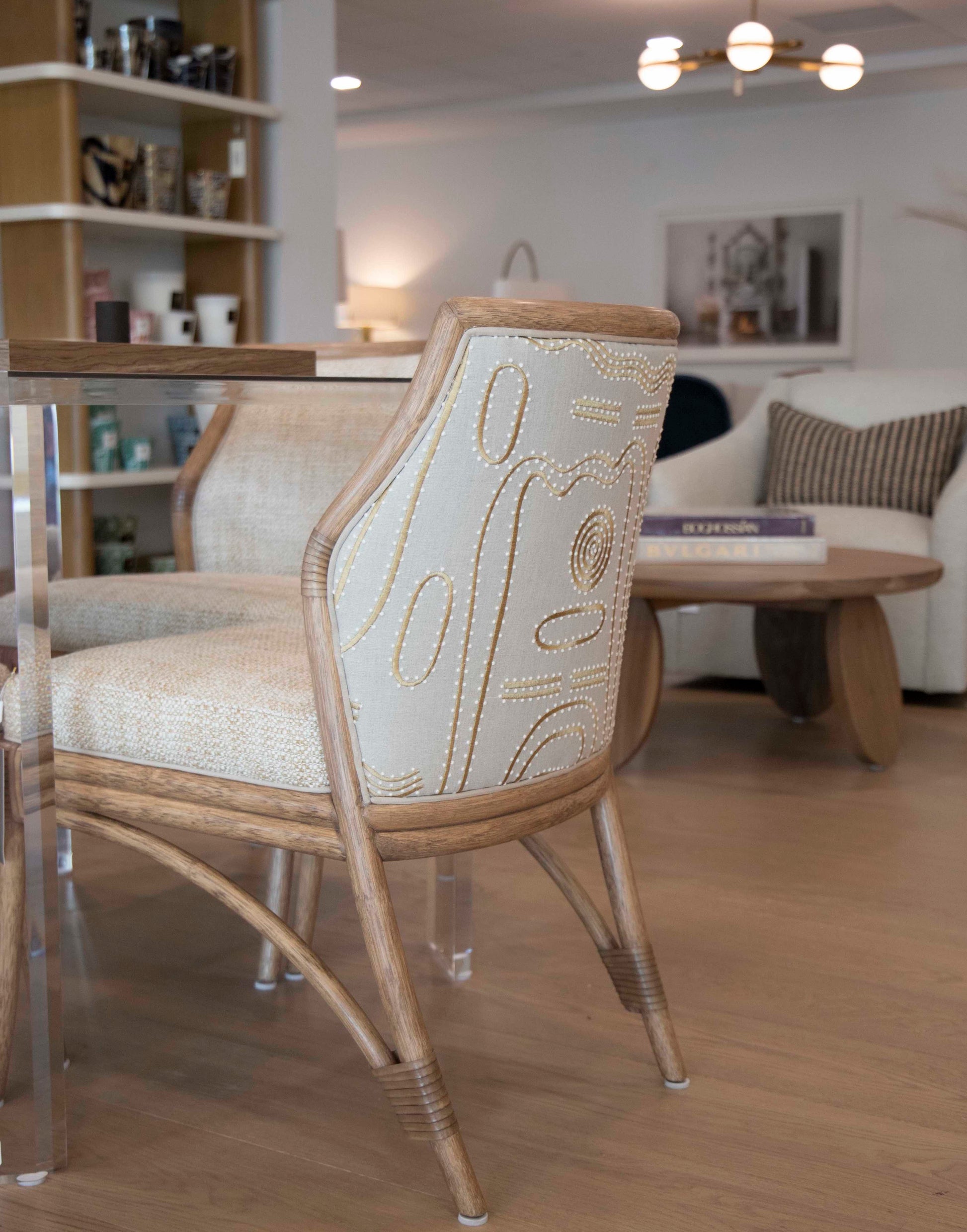 A modern Lantana Side Chair with wooden legs and patterned upholstery stands next to a glass table in a cozy living space, showcasing Lantana Side Chair craftsmanship. In the background, you can see a sofa, coffee table, and bookshelves.