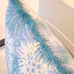 Close-up of the Bimini Sea Glass Outdoor Pillow, featuring a blue and white floral pattern and blue fringe, placed on a light-colored chair.