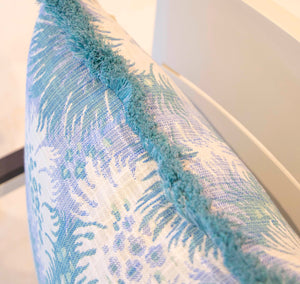 Close-up of the Bimini Sea Glass Outdoor Pillow, featuring a blue and white floral pattern and blue fringe, placed on a light-colored chair.