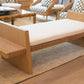 The Euclid Daybed, personalized with plush upholstery for extra comfort, showcases a refined wooden design with a cushioned seat. It is elegantly placed on a woven rug. Nearby, chairs adorned with patterned cushions complement a side table filled with books, crafting an inviting and cozy atmosphere.