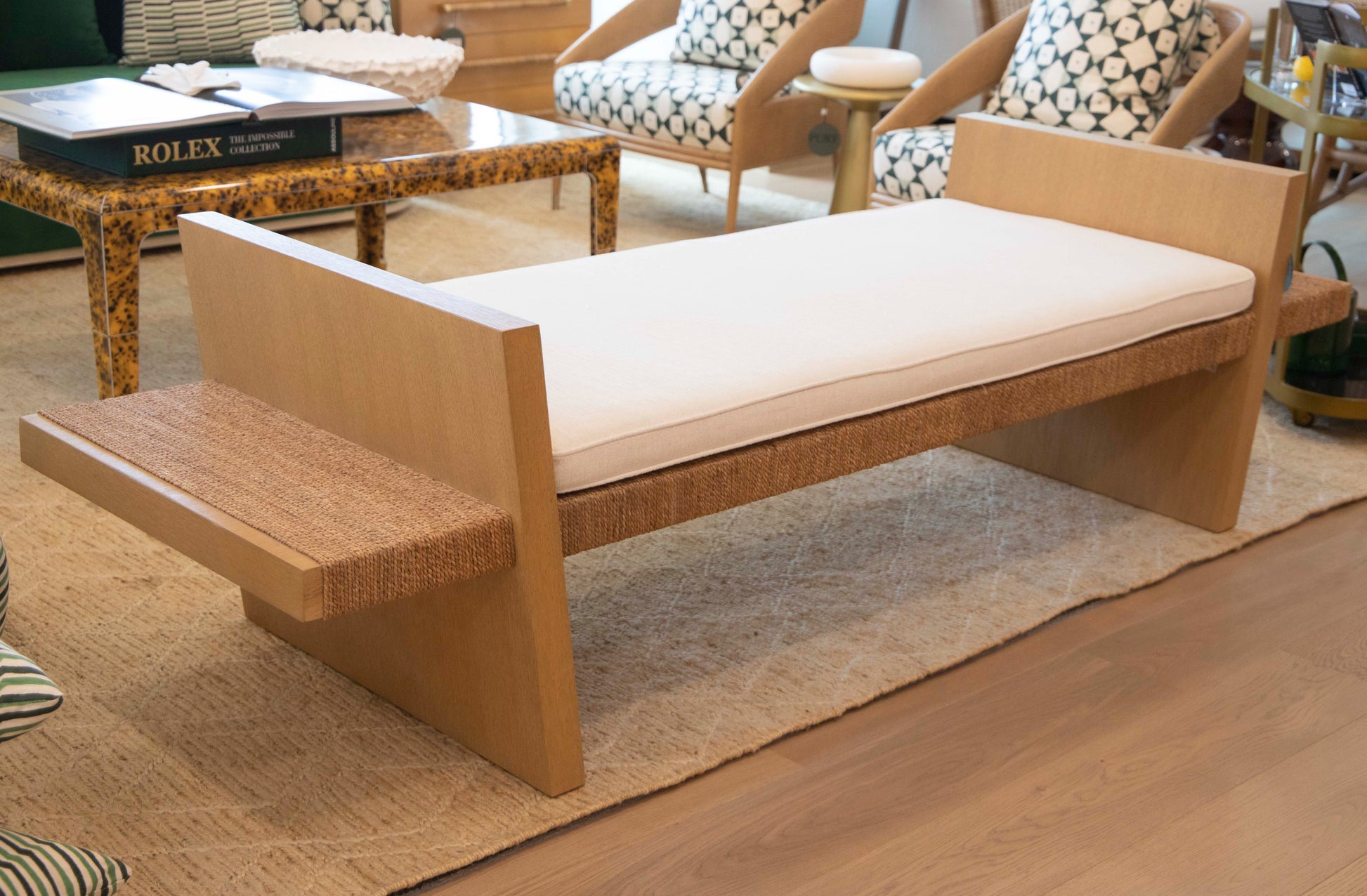 The Euclid Daybed, personalized with plush upholstery for extra comfort, showcases a refined wooden design with a cushioned seat. It is elegantly placed on a woven rug. Nearby, chairs adorned with patterned cushions complement a side table filled with books, crafting an inviting and cozy atmosphere.