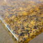 The image shows a close-up view of the Gerrit Cocktail Table's corner, showcasing a speckled, yellowish-brown surface with angular lines, a glossy finish, and exquisite metal inlays.