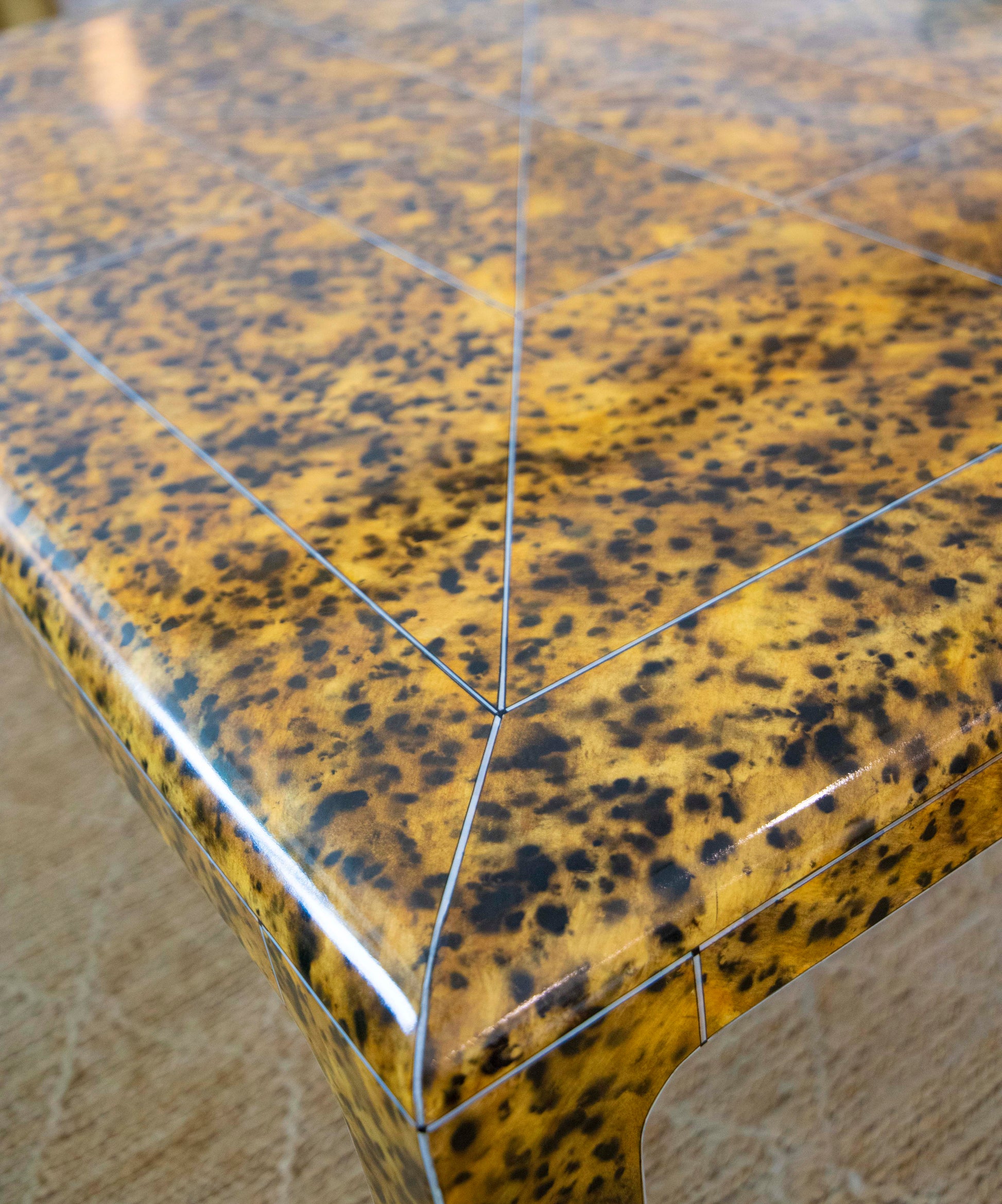 The image shows a close-up view of the Gerrit Cocktail Table's corner, showcasing a speckled, yellowish-brown surface with angular lines, a glossy finish, and exquisite metal inlays.