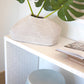 A white woven vase with large green leaves sits on a white table next to an opened book and a Porcelain Ribbed Garden Stool featuring a light blue glaze.