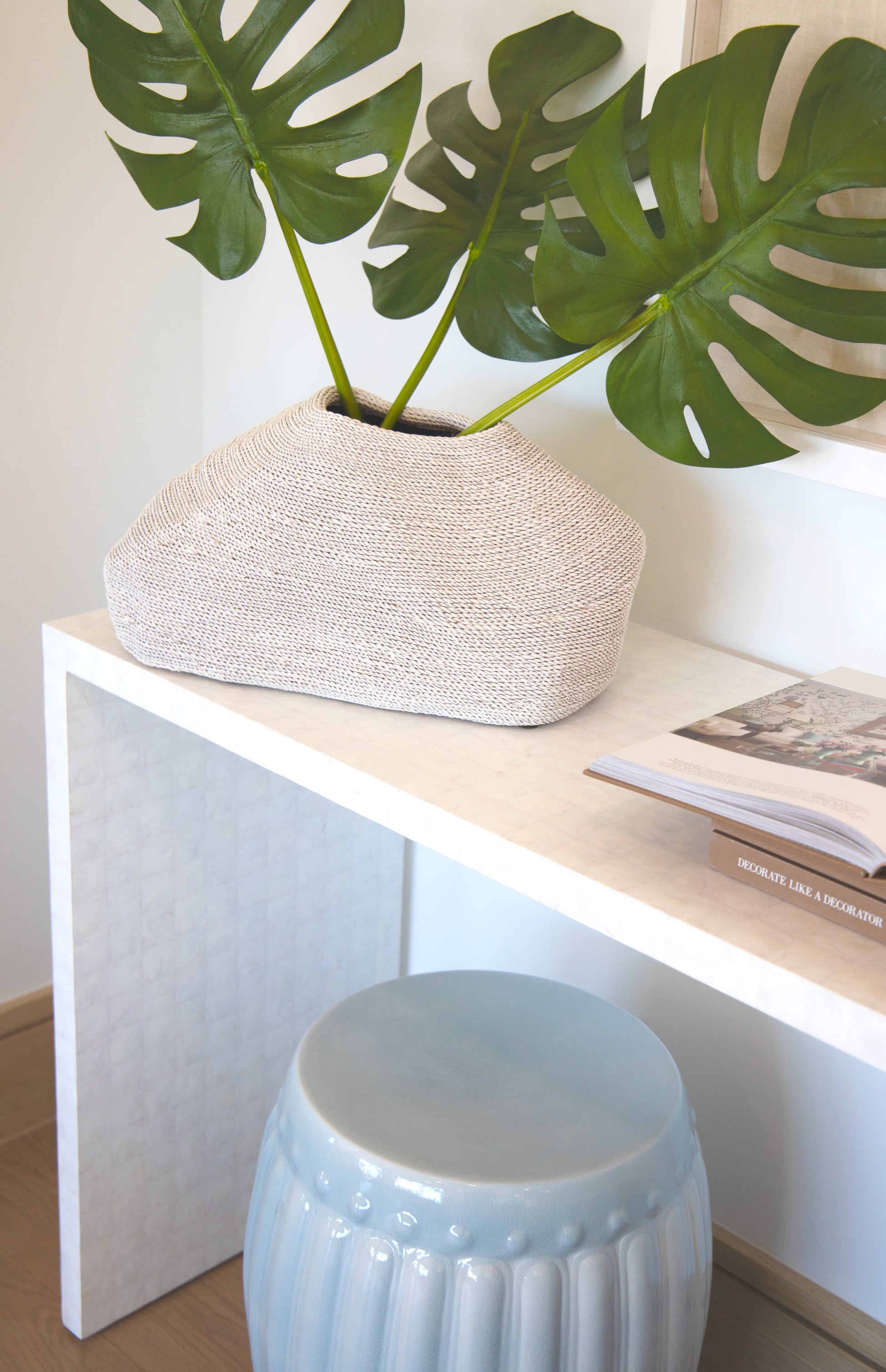 A white woven vase with large green leaves sits on a white table next to an opened book and a Porcelain Ribbed Garden Stool featuring a light blue glaze.