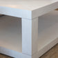 Close-up of the corner of a white wooden Silas Cocktail Table with an antiqued finish, showcasing its rustic texture. The clean lines highlight the lower shelf visible beneath the tabletop. A wooden tray is partially seen on the tabletop, adding a touch of charm.