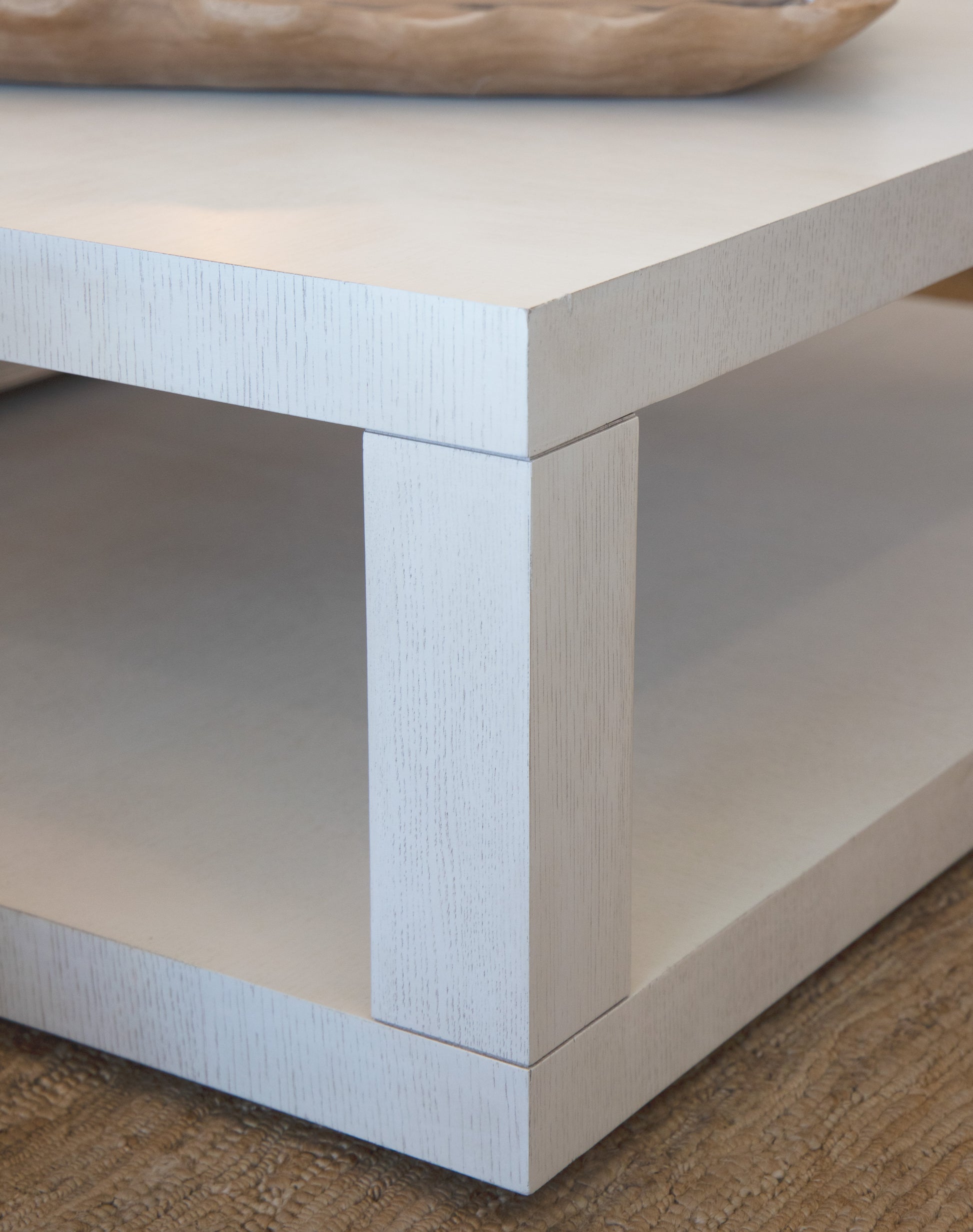 Close-up of the corner of a white wooden Silas Cocktail Table with an antiqued finish, showcasing its rustic texture. The clean lines highlight the lower shelf visible beneath the tabletop. A wooden tray is partially seen on the tabletop, adding a touch of charm.