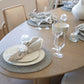 A neatly set Forum Dining Table with grey placemats, white plates, linen napkins, wine glasses, and a white centerpiece in a bright room evokes a Rome-inspired elegance through its classical design elements.