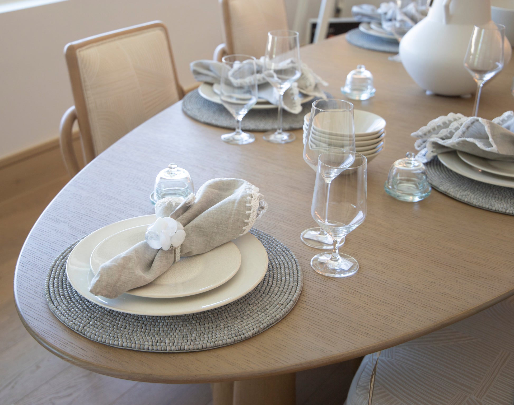 A neatly set Forum Dining Table with grey placemats, white plates, linen napkins, wine glasses, and a white centerpiece in a bright room evokes a Rome-inspired elegance through its classical design elements.