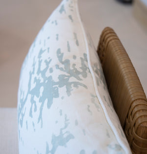 Close-up of a white cushion with a light blue abstract pattern, leaning against a wicker chair. Ripple Silver Jade Lumbar Pillow like this one is crafted with special attention to detail and uses the finest materials.