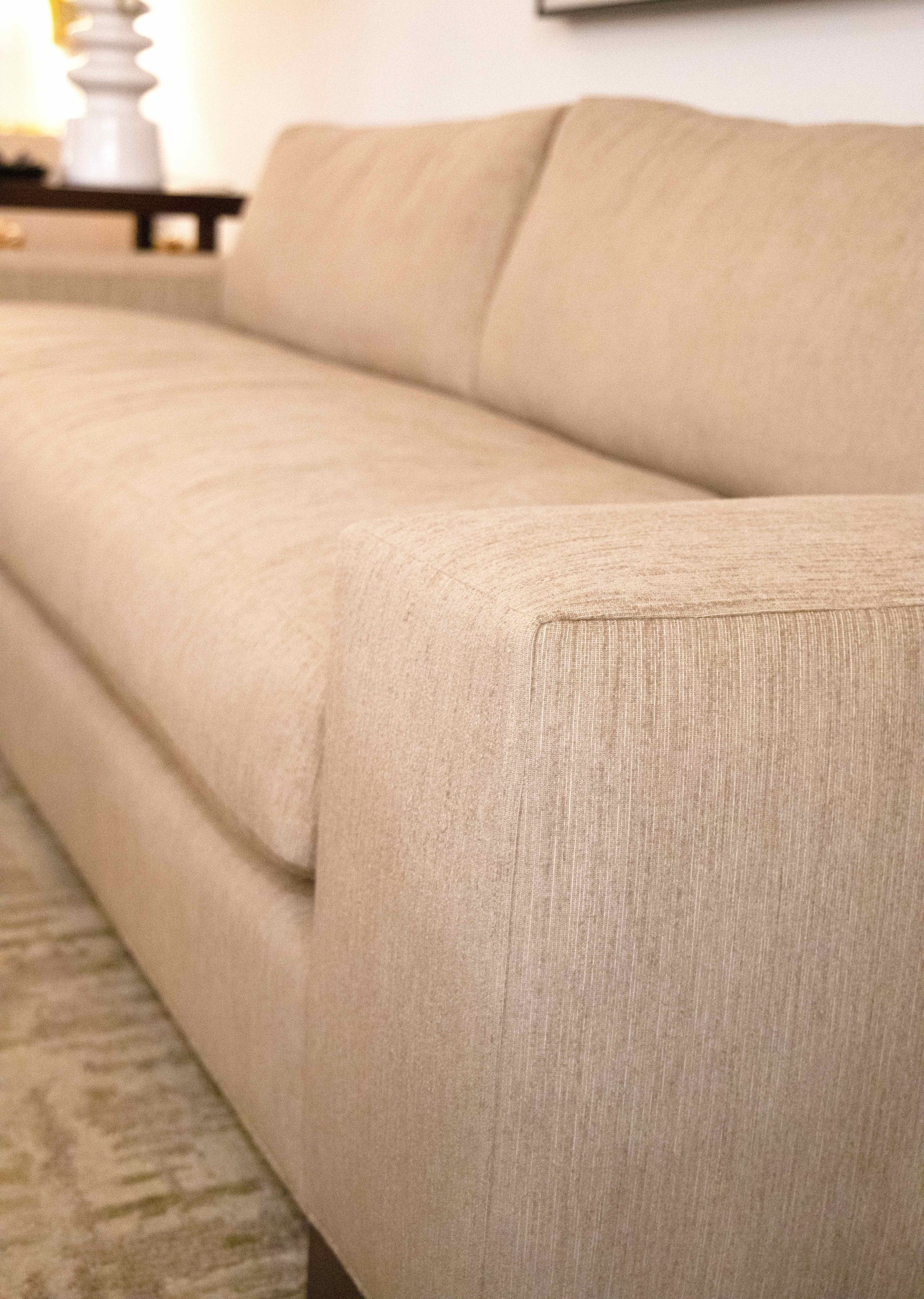 A close-up view of the Burke Sofa with a modern design, showcasing the texture of the upholstery and the armrest detail. A lamp and a side table are visible in the background, adding a touch of classic simplicity to the industrial elements.