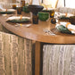 An Ellipse Dining Table set for six with green and brown tableware, surrounded by patterned cushioned chairs, and a plant in the background showcases refined textures.