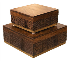 Two square, wooden boxes from the Oti Brown Box Collection with carved symbols and geometric patterns on their sides are stacked on top of each other. Crafted from mango wood, the surface of the boxes is smooth and polished.