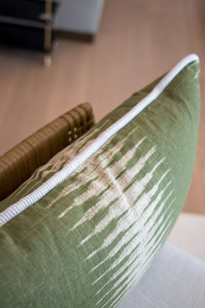 Close-up of the Ikat Olive Lumbar Pillow—a custom-made green decorative pillow with a white abstract pattern and white piping—resting on a chair with a wooden armrest.