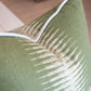 Close-up of the Ikat Olive Pillow, a green decorative cushion featuring white abstract streak patterns and white piping along the edges, resting on a beige surface.