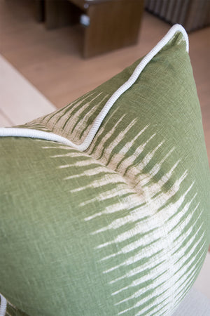 Close-up of the Ikat Olive Pillow, a green decorative cushion featuring white abstract streak patterns and white piping along the edges, resting on a beige surface.