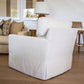 The Blair Swivel Chair, featuring white upholstery, a modern square back, and a skirted base, sits on a wooden floor in a living room with a beige sofa, pillows, lamp, and wooden chair in the background.