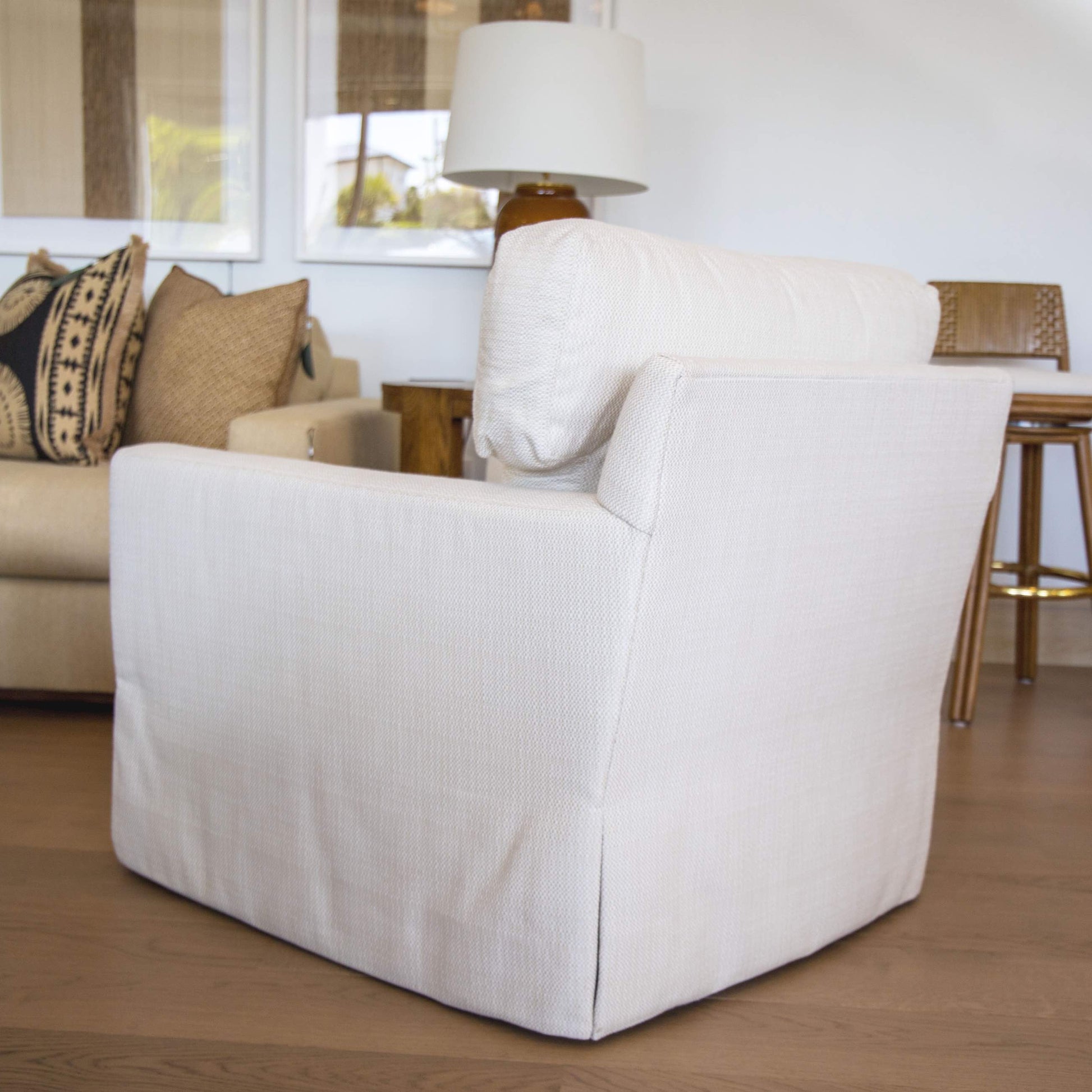 The Blair Swivel Chair, featuring white upholstery, a modern square back, and a skirted base, sits on a wooden floor in a living room with a beige sofa, pillows, lamp, and wooden chair in the background.