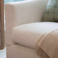 Close-up of the Barrel Swivel Chair with custom white upholstery, rounded back, a green patterned pillow, and a beige fringed throw draped over the seat, placed beside a wooden side table.