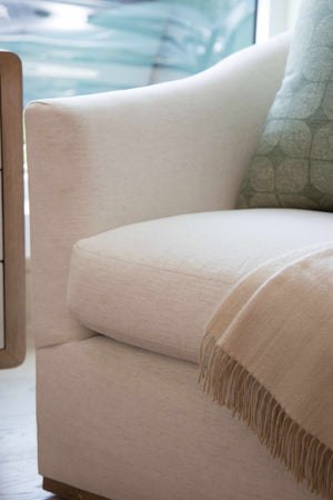 Close-up of the Barrel Swivel Chair with custom white upholstery, rounded back, a green patterned pillow, and a beige fringed throw draped over the seat, placed beside a wooden side table.