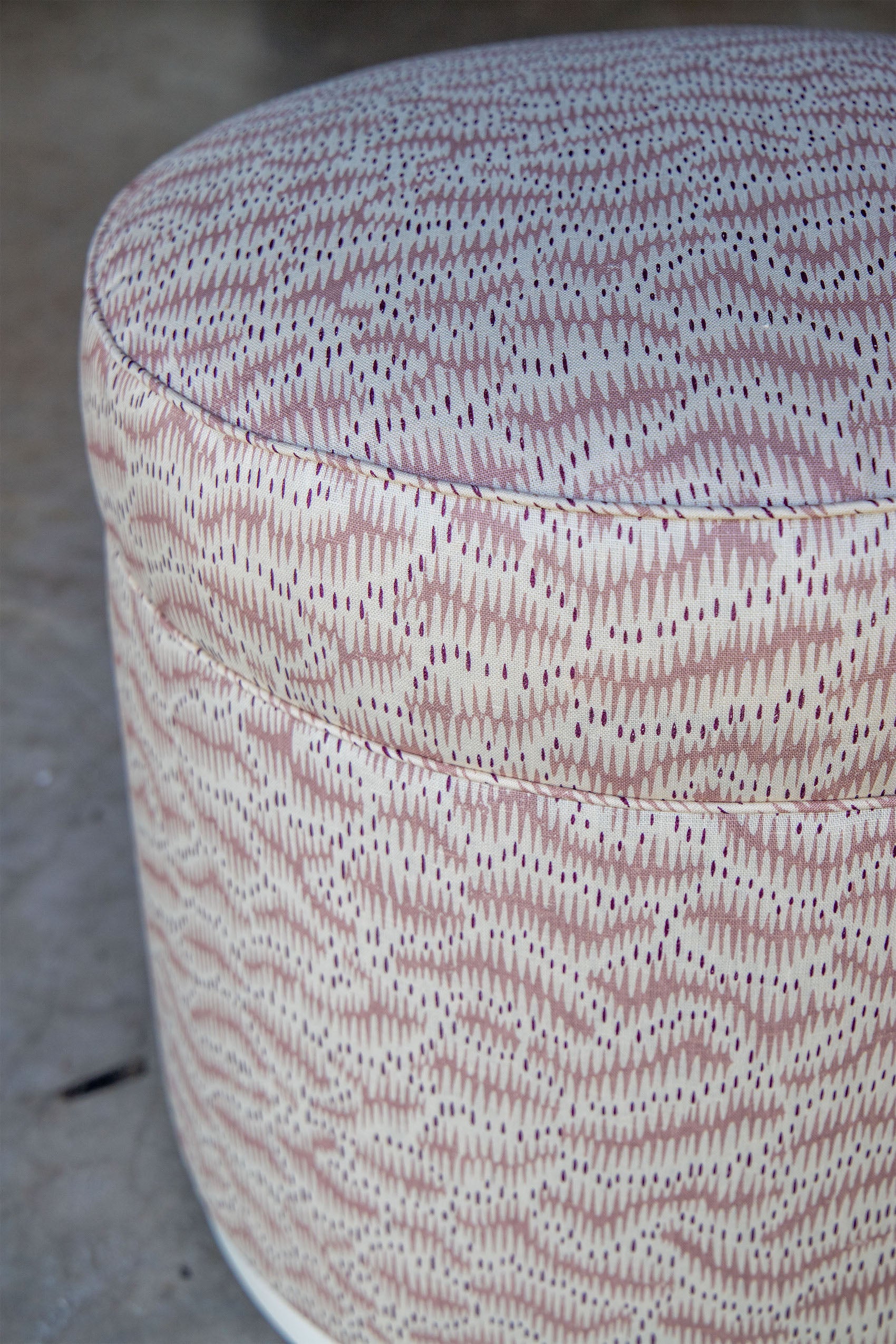 The Grant Swivel Ottoman, upholstered in Daphne Feathers Rose beige fabric with a mauve and white geometric pattern, adds charm to elegant living spaces. Shown here on a concrete floor.