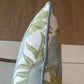A close-up of the Carnival Celeste Outdoor Pillow, featuring a floral design and blue trim, displayed vertically on a chair with tile and wood floor background.