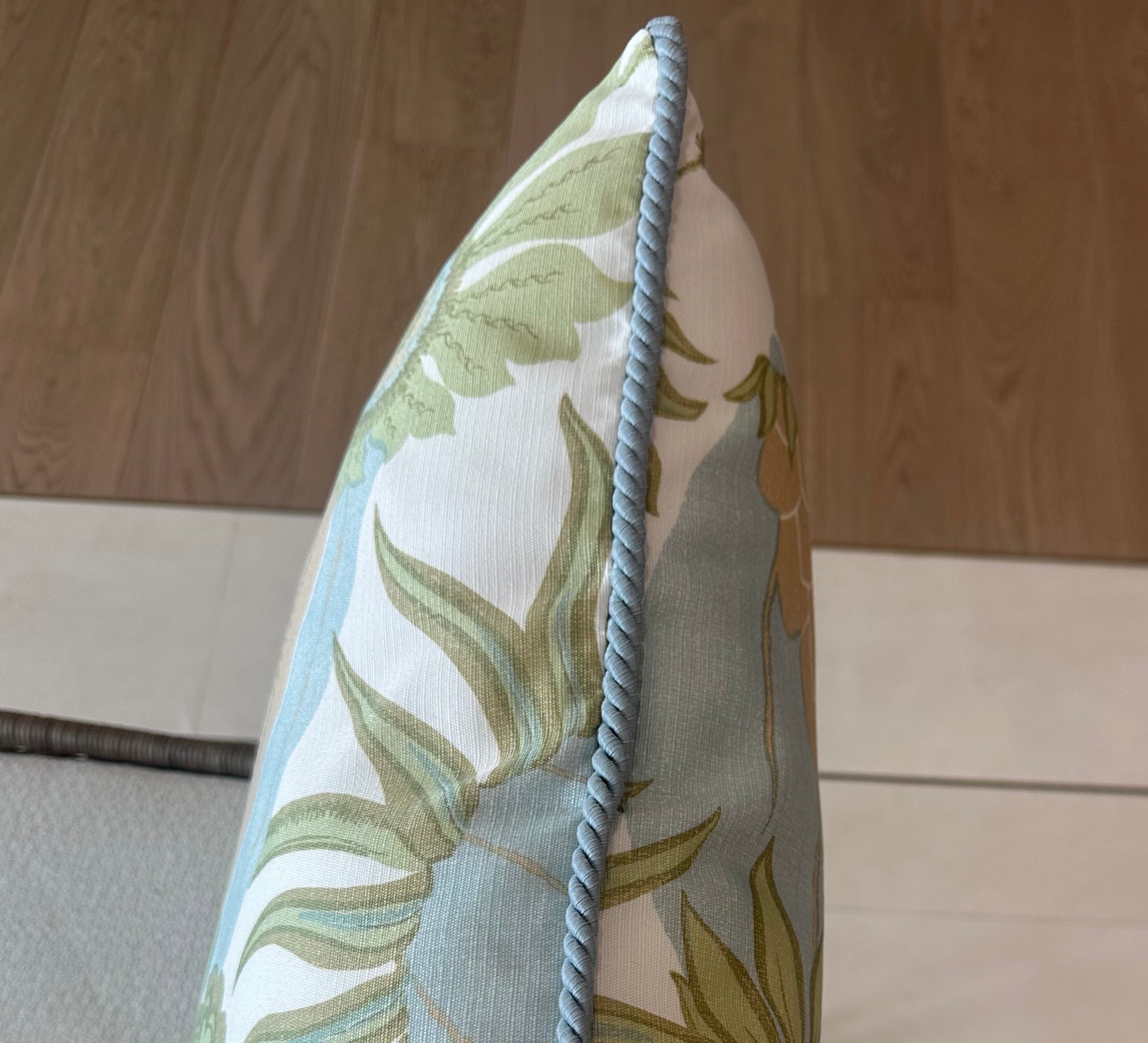 A close-up of the Carnival Celeste Outdoor Pillow, featuring a floral design and blue trim, displayed vertically on a chair with tile and wood floor background.