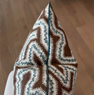 A close-up of the Moorish Maze Pillow, featuring a custom brown and white pattern with a pointed edge, rests on a light surface and wooden floor, highlighting the premium materials used for this unique cushion.