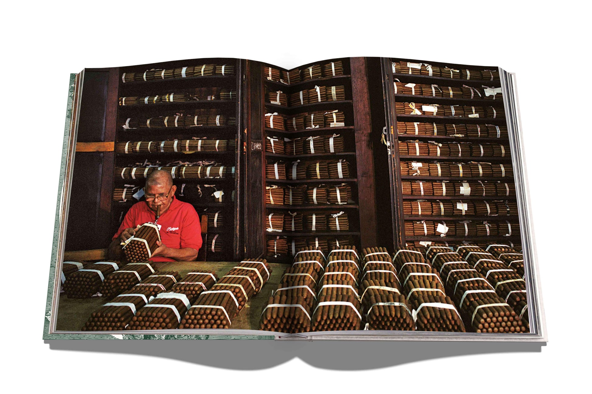 A man in a red shirt inspects bunches of hand-rolled cigars in a room with shelves full of cigar bundles. The scene is displayed across the pages of an open book, part of The Impossible Collection of Cigars.