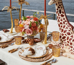 A waterfront table set with gold and bamboo decor features the Kim Seybert Royal Palm Placemat, adorned with hand-applied beads for a tropical touch, along with gold palm tree candle holders, floral centerpiece, gold-rimmed glasses, and a giraffe sculpture.