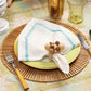 A table setting features the Kim Seybert Spoke Placemat, a yellow plate, a white napkin with blue trim and decorative ring, fork, knife, glassware, all atop a geometric-patterned tablecloth.
