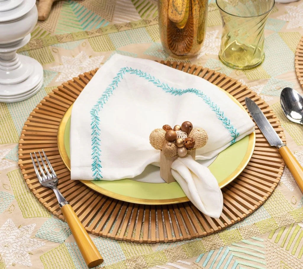 A table setting features the Kim Seybert Spoke Placemat, a yellow plate, a white napkin with blue trim and decorative ring, fork, knife, glassware, all atop a geometric-patterned tablecloth.